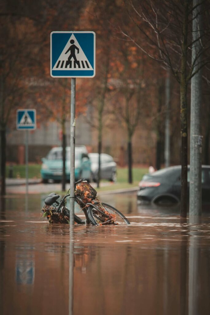 "An urban street submerged after a flood, with a bicycle leaning against a pedestrian crossing sign, partially underwater and tangled with leaves. The image highlights the disruptions a single flood can cause, from damaged transportation infrastructure to blocked pathways for pedestrians and vehicles. In the background, partially flooded cars and autumn trees emphasize the widespread impact on everyday urban life, illustrating the need for resilient urban planning and climate impact chains (CICs) to anticipate and mitigate such cascading effects."